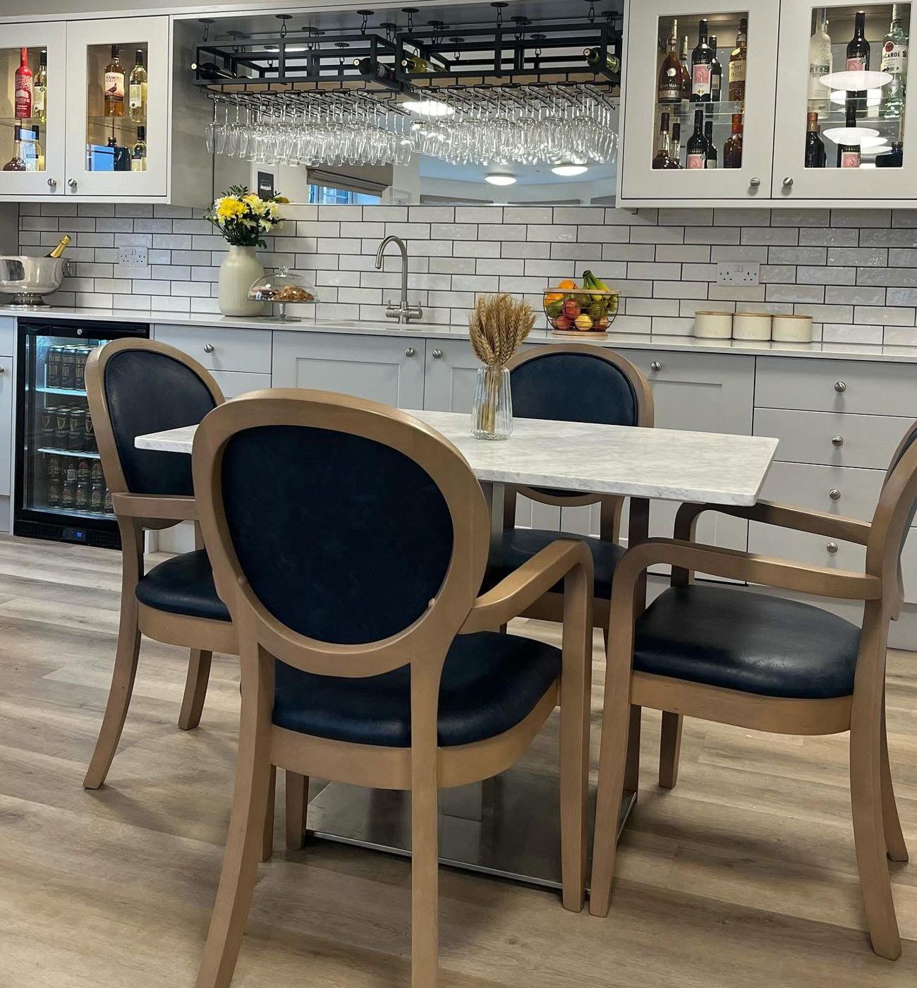 A dining table with blue upholstered chairs, laid with a vase of dried wheat