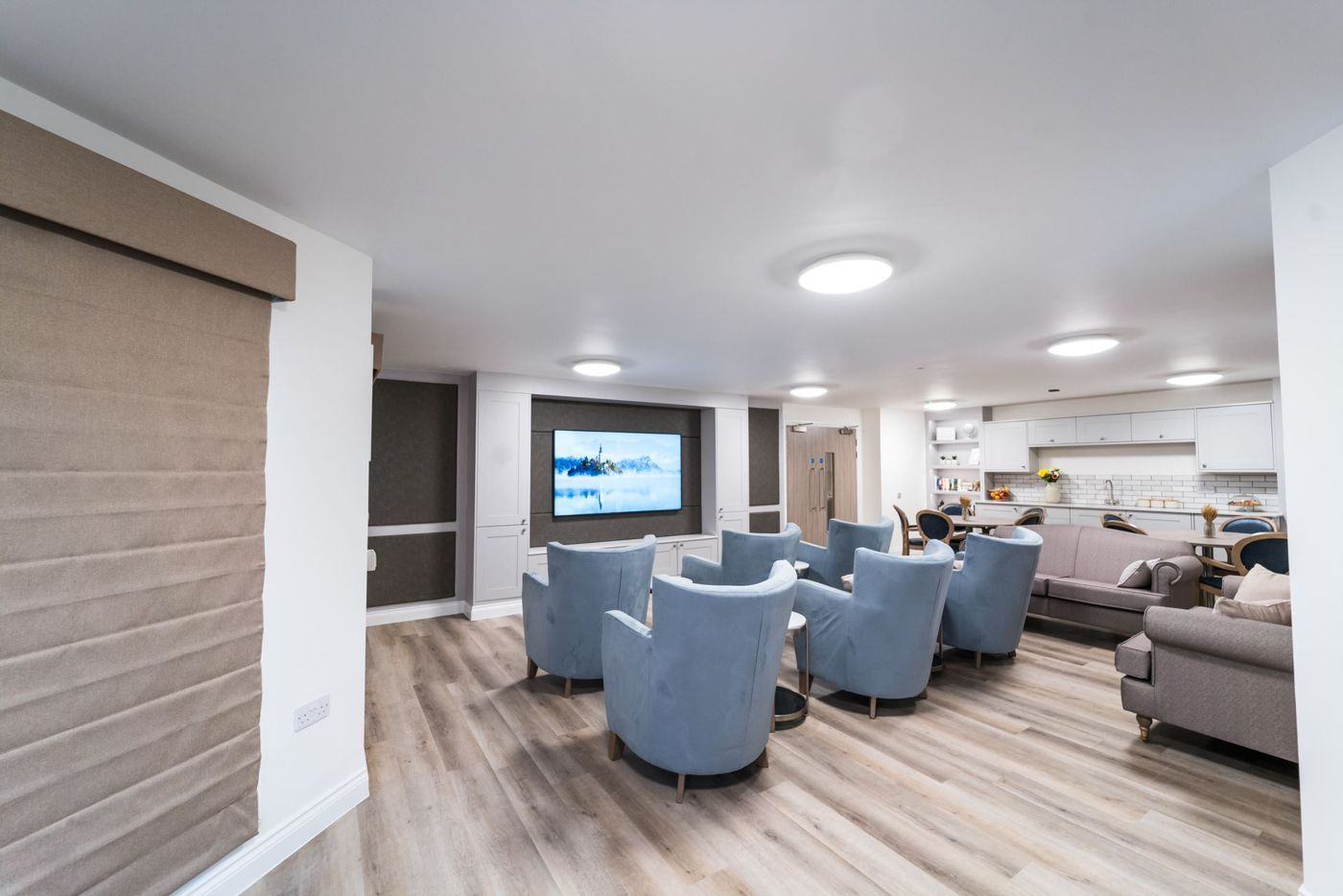 A lounge with armchairs facing a large screen, and a kitchenette