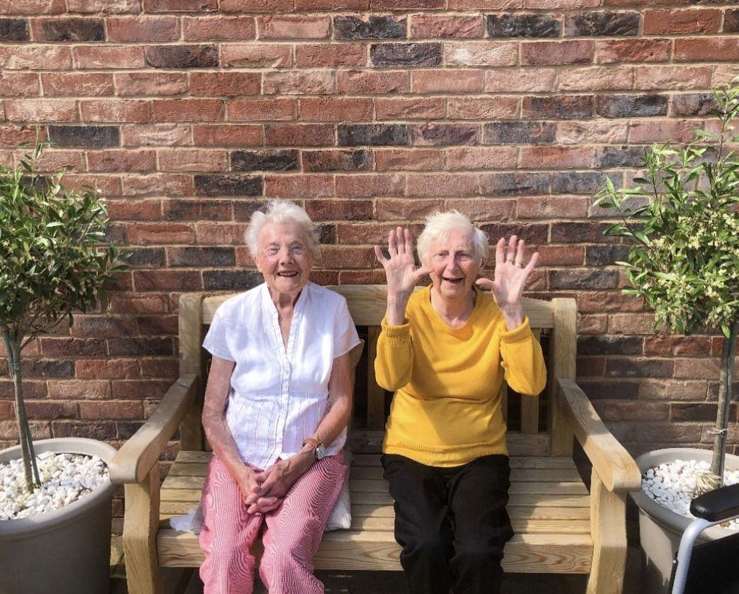 Two smiling residents waving from a garden bench at Hampton Manor